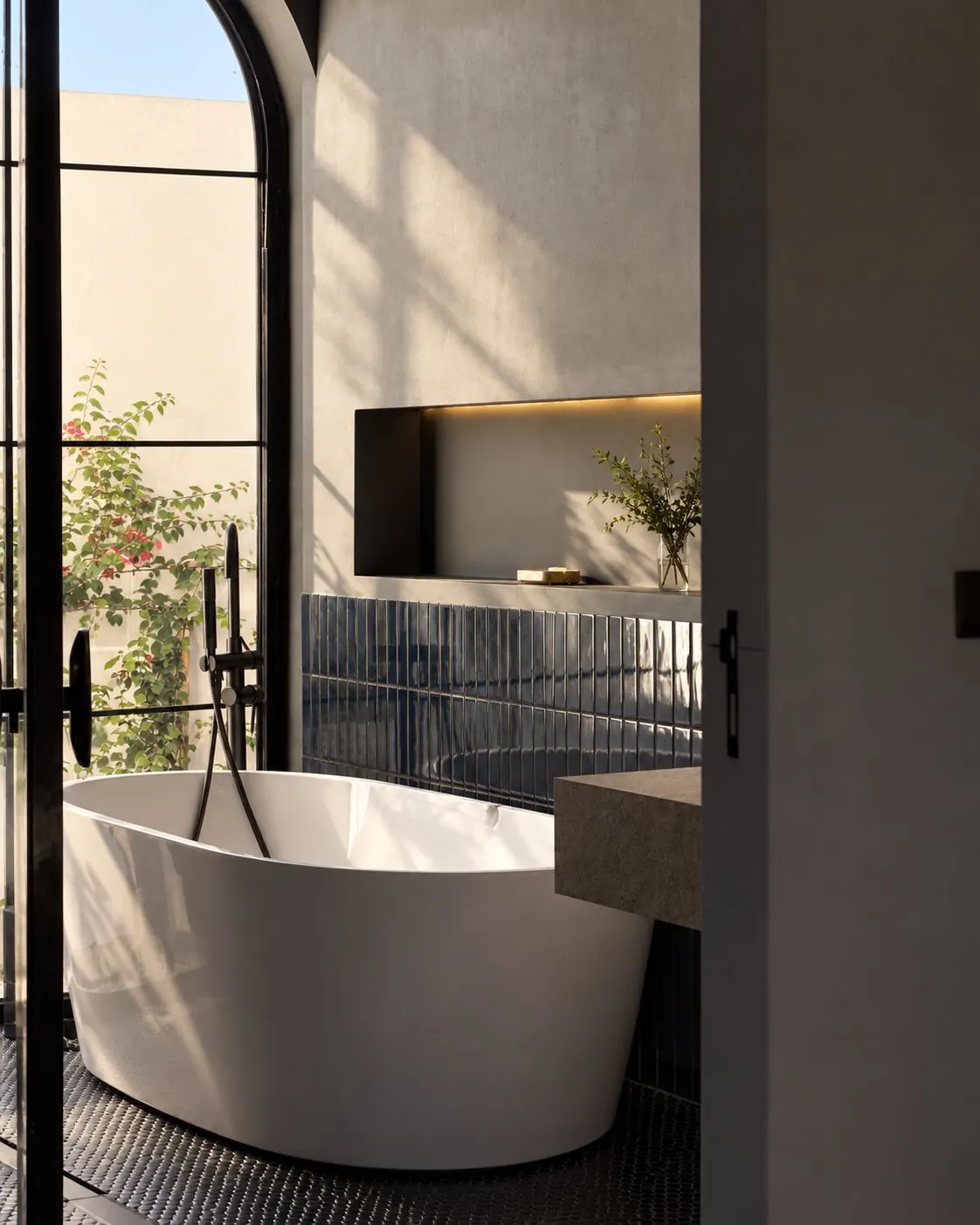 Freestanding white bath by an arched steel window, dark blue glazed tiles and a black floor-standing mixer