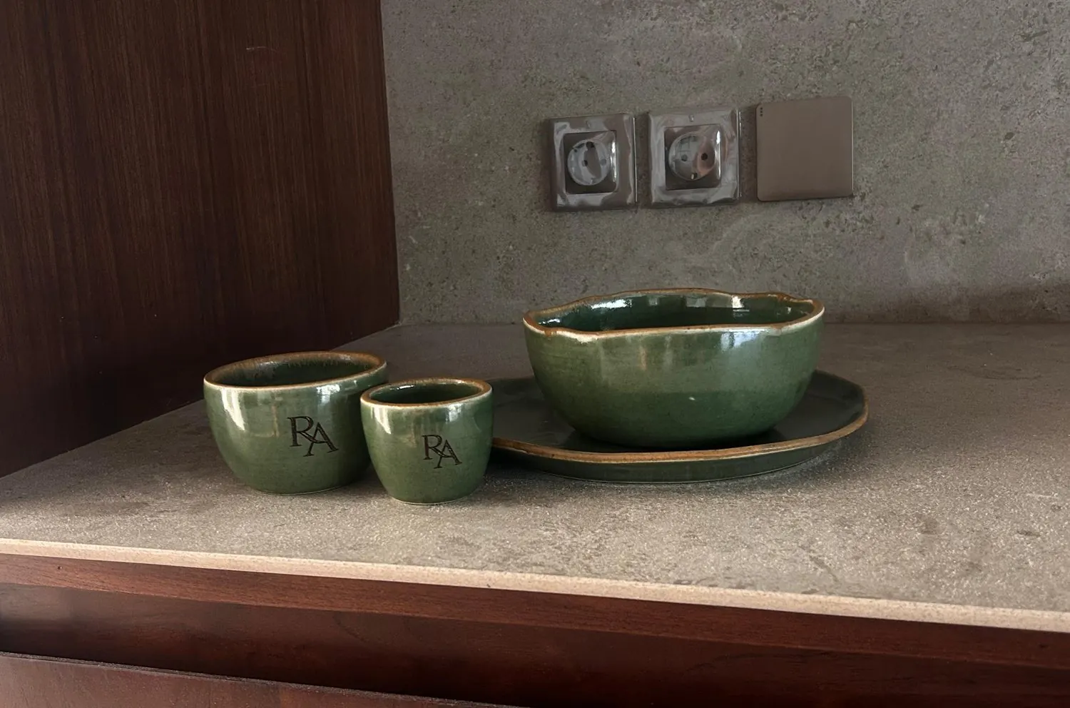 Green glazed cups and a bowl with the RA monogram on a stone worktop against a walnut cabinet