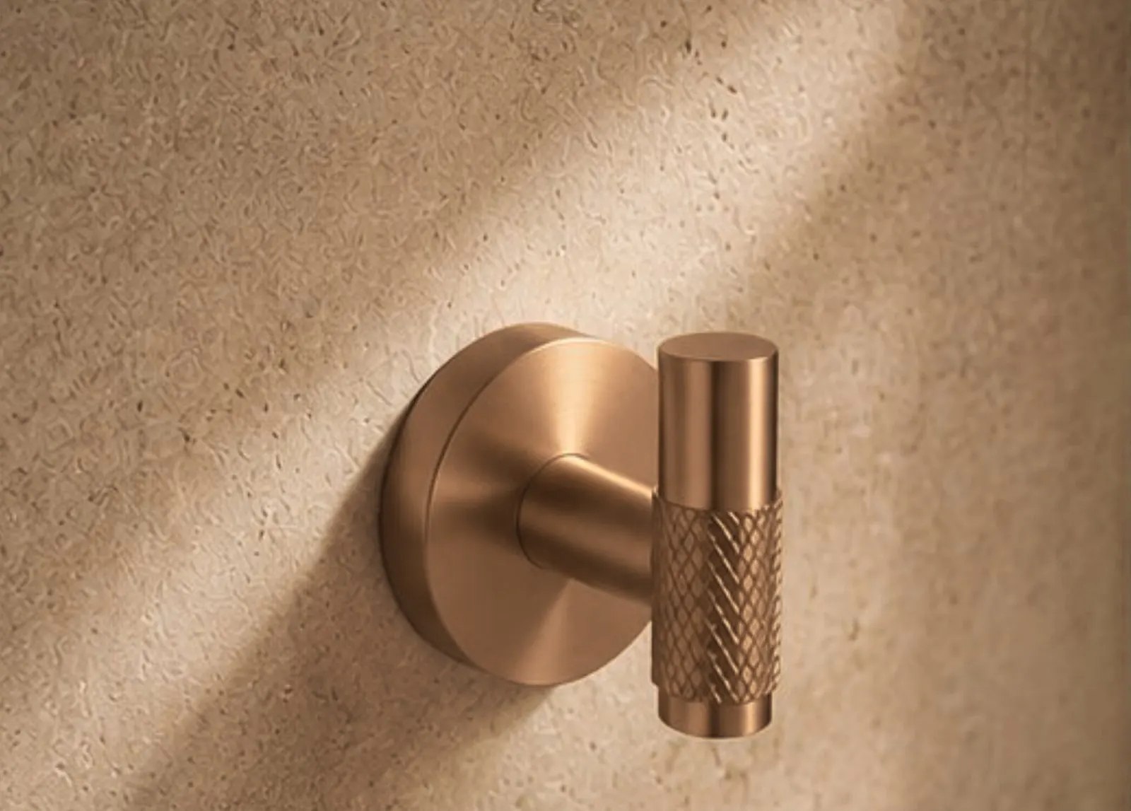 Close-up of a rose gold robe hook with a diamond-knurled grip, sunlight across a stone wall